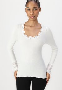 Haut à manches longues blanc à côtes, présentant un col en feston et des accents en dentelle aux poignets. Texture douce avec un design ajusté.