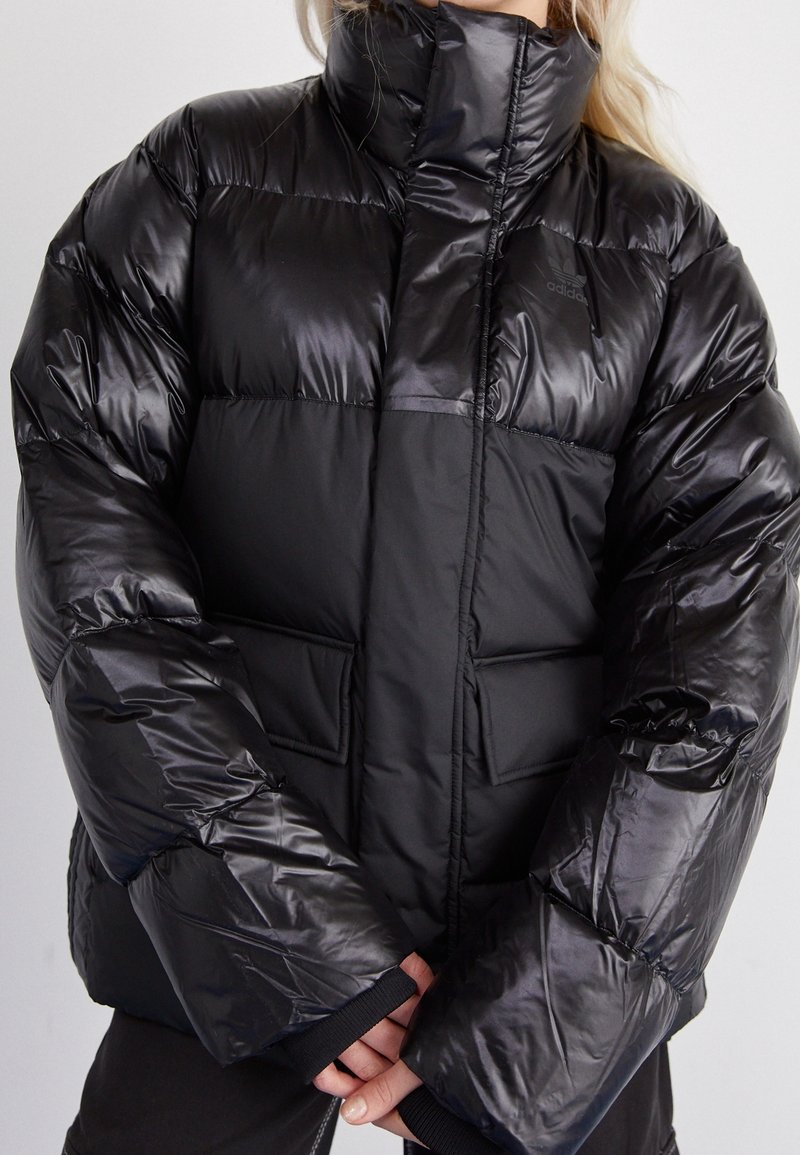 Black puffer jacket with a high collar, shiny texture, padded sections, and front pockets. Features elastic cuffs and subtle branding.