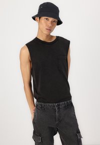 Zwarte mouwloze T-shirt met een borstzak, gecombineerd met donkergrijze cargobroek. Model draagt een zwarte bucket hat. Eenvoudig, casual ontwerp.