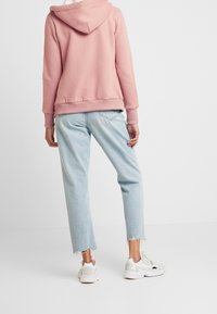 Persona con una sudadera rosa, pantalones vaqueros de dobladillo deshilachado en un azul claro, y zapatillas blancas, de pie contra un fondo blanco liso.