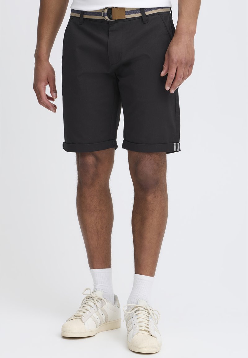 Shorts noirs avec un ourlet replié, en tissu lisse. Dotés de poches latérales et d'une ceinture à rayures brunes. Portés avec des baskets blanches et des chaussettes.