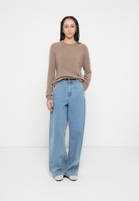 Καφέ πλεκτό πουλόβερ με στρογγυλή λαιμόκοψη, σε συνδυασμό με ψηλόμεσα, φαρδιά μπλε τζιν παντελόνια. Το μοντέλο στέκεται με τα χέρια στα πλάγια.