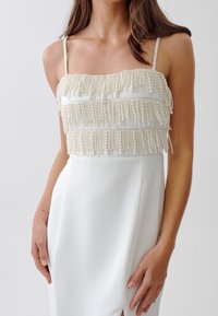 Bílé šaty s tiered perlou zdobeným korzetem, tenkými ramínky a elegantním siluetou s bočním rozparkem.