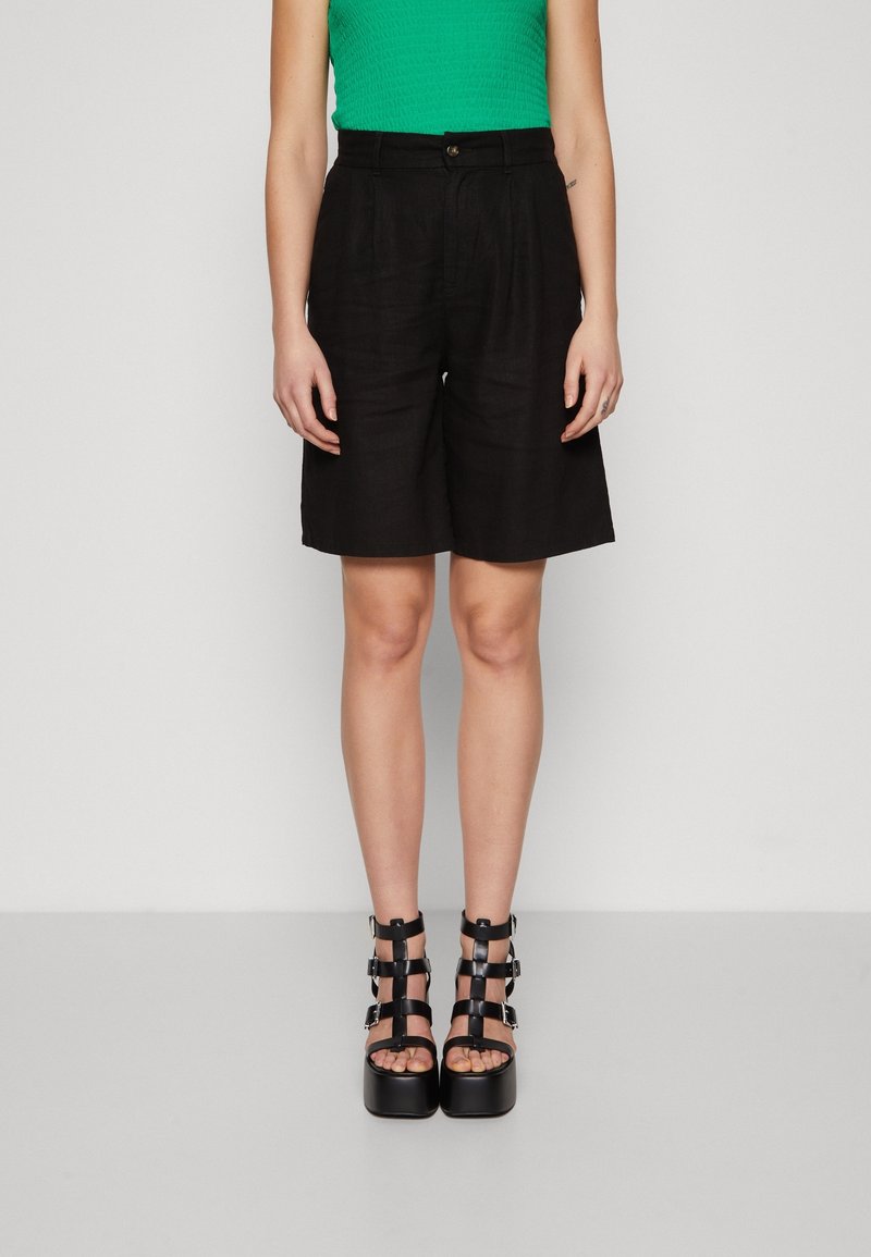 Personne portant un short taille haute noir, un haut sans manches vert et des sandales à plateforme noires à lanières, se tenant contre un fond neutre.