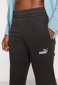 Μαύρο παντελόνι φόρμας με ελαστική μέση, που φέρει λευκό λογότυπο Puma στο αριστερό μηρό και έχει λεία υφή. Φορεμένο με ανοιχτό μπλε μπουφάν με φερμουάρ.