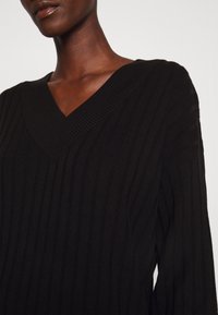 Personne à la peau foncée portant un pull noir en maille côtelée avec un décolleté en V profond sur un fond clair.