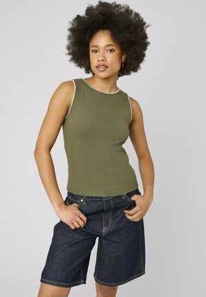 Femme avec coiffure afro portant un top côtelé sans manches vert olive et un short en jean foncé longueur genou, debout les mains dans les poches.