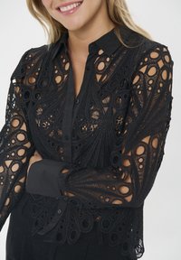 Black blouse featuring intricate lace detailing with circular cutouts and a classic collar, buttoned front, and long, sheer sleeves.