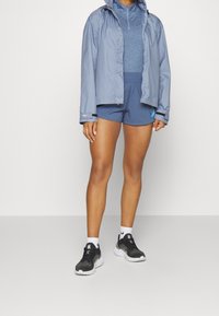 Persona vestida con una chaqueta azul clara, una camiseta deportiva azul, pantalones cortos atléticos azules, calcetines blancos y zapatillas de correr negras, de pie sobre un suelo gris.