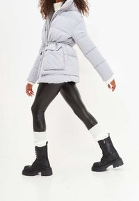 Lichtgrijze donsjas met een tailleband, gecombineerd met zwarte legging van kunstleer en zwarte combatlaarzen met witte gebreide manchet.