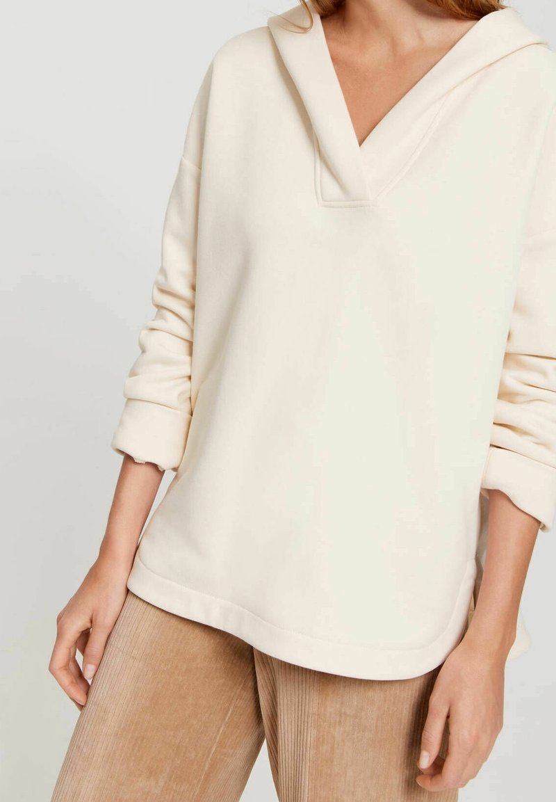 Krémová mikina s kapucí, s výstřihem do V, volným střihem a ohrnutými rukávy. Materiál vypadá měkce a má strukturu, ideální pro každodenní nošení.