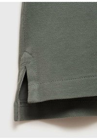 Šedozelená polo košile s texturovanou, vaflovou tkaninou; má boční rozparky na lemu a zesílené švy kolem okrajů.