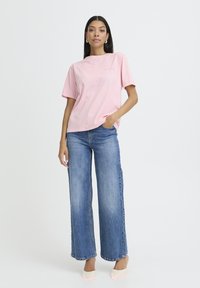 Frau steht mit einer Hand in der Tasche, trägt ein hellrosa Kurzarmshirt, blaue weite Jeans und weiße spitze Schuhe.