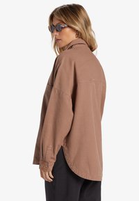 Lichtbruine oversized shirt met lange mouwen en knoopmanchetten, voorzien van een gebogen zoom en een klein logo-detail op de rug.