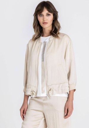 Femme aux cheveux bruns ondulés portant une veste beige légère sur un haut blanc avec un détail métallique vertical, assortie à un pantalon beige.