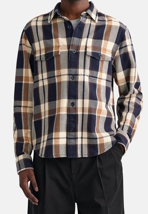 Mężczyzna ubrany w granatową, brązową i kremową koszulę w kratę z kieszeniami na piersi oraz czarne spodnie na białym tle.