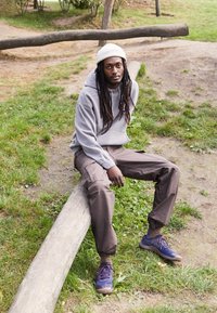 Felpa grigia, pantaloni cargo marroni e sneaker blu. Un uomo con lunghi capelli neri è seduto su un tronco in un'area erbosa circondata da alberi.