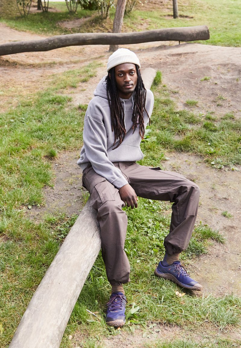 Felpa grigia, pantaloni cargo marroni e sneaker blu. Un uomo con lunghi capelli neri è seduto su un tronco in un'area erbosa circondata da alberi.