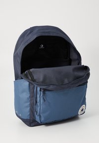 Marineblå nylon ryggsekk med glidelås foran lomme, sirkulært logo-merke og mesh-fôret innside. Glatt tekstur og moderne design.