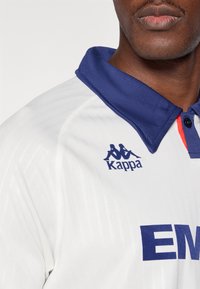 Biele športové tričko s námornícko modrým golierom, na ktorom je modré logo Kappa a tučný modrý nápis "EM". Jemná textúra a dizajnové detaily sú viditeľné.