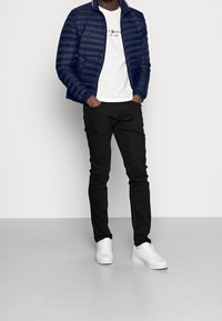 Man som bär en marinblå puffjacka över en vit t-shirt med logga, svarta jeans och vita sneakers, står med händerna i fickorna framför en neutral bakgrund.