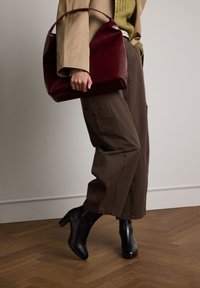 Bordeauxrote Lederhandtasche mit strukturiertem Design, kombiniert mit braunen, weit geschnittenen Cargo-Hosen und schwarzen, Absatz-ankle Boots auf einem Holzboden.