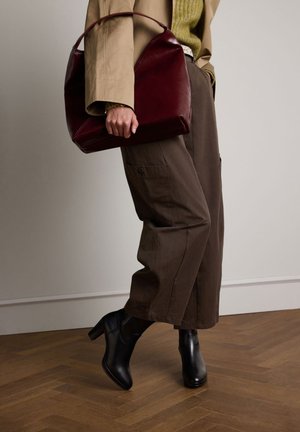 Maroen leren handtas met een gestructureerd ontwerp, gecombineerd met bruine cargo-broeken met wijd uitlopende pijpen en zwarte enkellaarsjes met hak op een houten vloer.