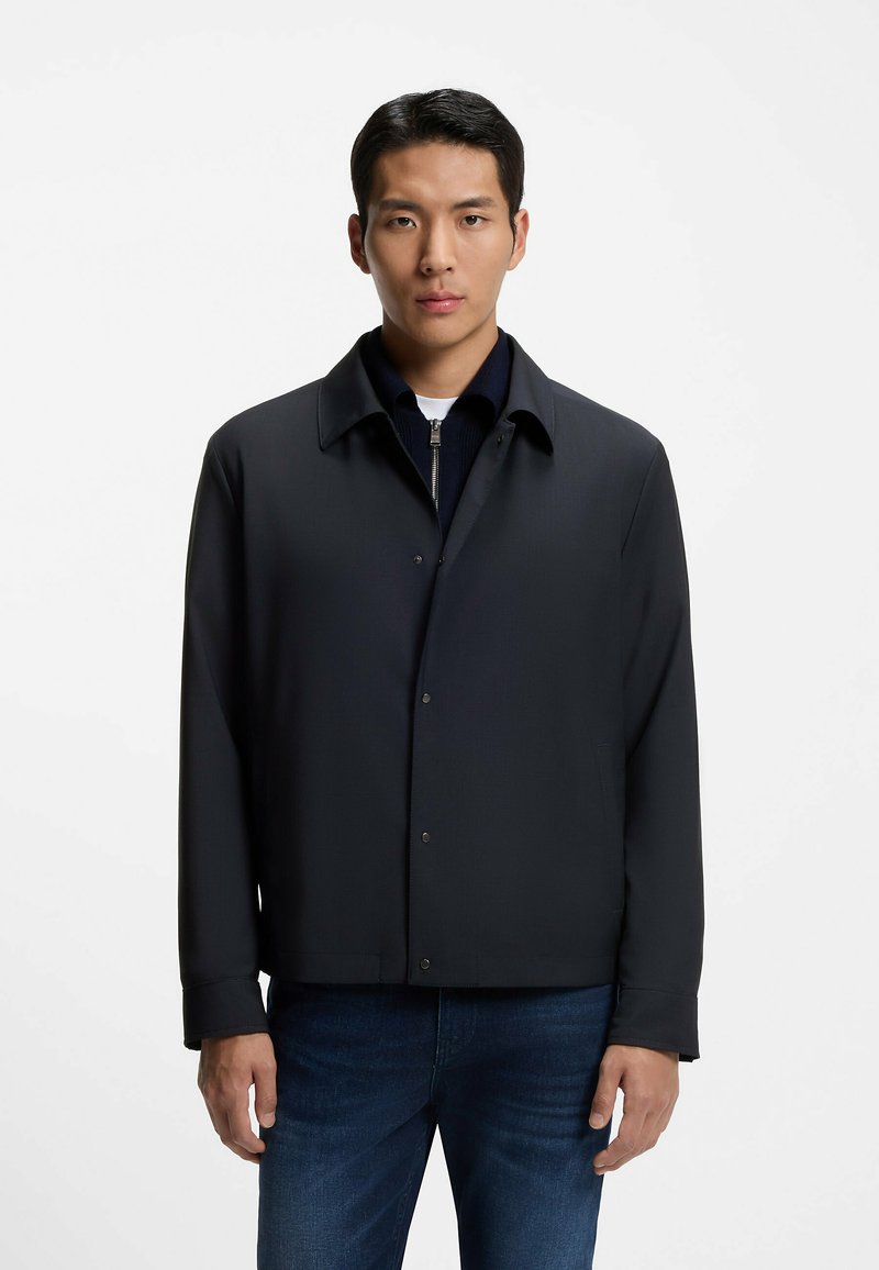 Dark blue jacket featuring a collared neckline, snap front closure, and long sleeves. Smooth fabric with a structured, modern fit.