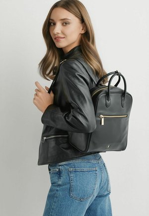 Młoda kobieta o jasnobrązowych włosach, ubrana w czarną skórzaną kurtkę i niebieskie jeansy, niosąca na jednym ramieniu czarny skórzany plecak.