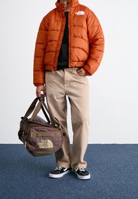 Orange puffer jacket, black shirt, beige pants, carrying a brown duffle bag with yellow logo. Black sneakers with white soles.