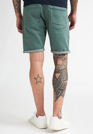 Shorts en coton vert avec revers roulés, dotés de poches arrière et d'une texture lisse. Tatouages remarquables sur les jambes, visibles au niveau des mollets.