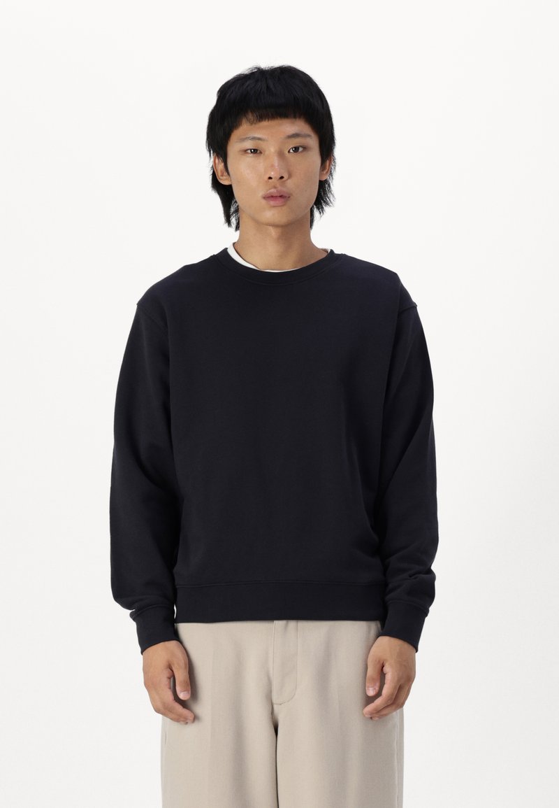 Sort bomulds sweatshirt med rund hals og dropped shoulders, parret med lys beige skræddersyede bukser. Minimalistisk design, ingen mønstre.
