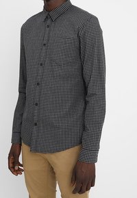 Camisa de manga larga a cuadros en negro y gris con cuello abotonado, un bolsillo en el pecho y un diseño ajustado. La textura de la tela parece suave.