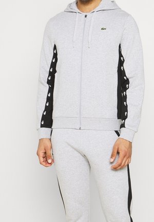 Mann trägt hellgrauen Kapuzenpullover mit Reißverschluss und passende Jogginghose mit schwarzen Seitenstreifen und weißen Logos auf Ärmeln und Hosen.