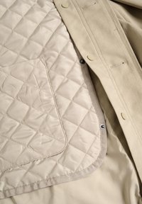 Beige ytterplagg med en quiltad innerfoder. Detaljer inkluderar en ficka med patch, tryckknappar och en slät textur med en kontrasterande fåll.