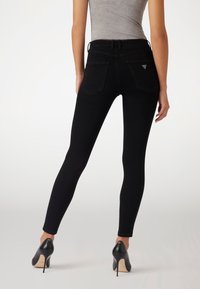 Black skinny jeans made of denim, featuring a high waist, two back pockets, and subtle branding on the right pocket. Worn with black high heels.