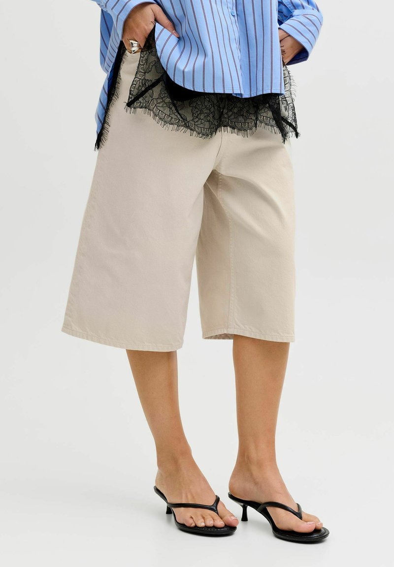 Persona vistiendo pantalones beige de pierna ancha y cortos, blusa de encaje negra bajo camisa a rayas azul, y sandalias negras de tacón, de pie sobre un suelo blanco.