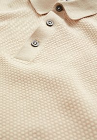 Beige textured polo shirt with a collared neckline, featuring three dark gray buttons and a pocket on the left side.