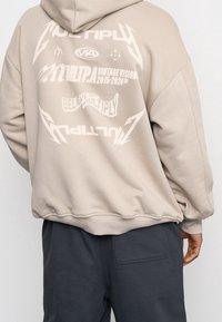 Sweat à capuche beige clair avec un grand texte graphique blanc au dos, comprenant des slogans et des logos. Tissu texturé et coupe décontractée visibles.