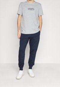 Camiseta de algodón gris con un logo azul y rojo que dice "HILFIGER EST. 1985", combinada con joggers azul marino y zapatillas blancas. Corte holgado, diseño casual.
