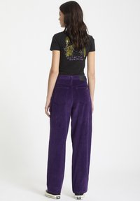 Pantalon large en velours côtelé violet avec des côtes verticales distinctives, muni de poches arrière et d'une étiquette à la taille, associé à un t-shirt graphique noir.