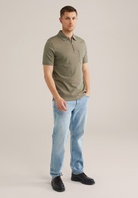 Polo de manga corta color verde oliva con cuello con cremallera y bolsillo en el pecho izquierdo, combinado con jeans de mezclilla azul claro y zapatos negros.