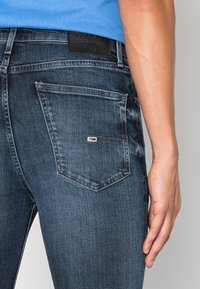 Mörka denimjeans med slim fit, med en distinkt bakficka och en svart varumärkesetikett på midjan. Slät textur.