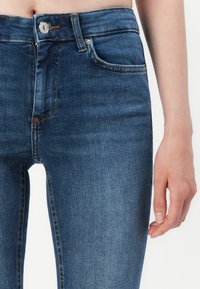 Tunna blå denimjeans med hög midja, med dragkedja, framfickor och kontrasterande sömmar. Slät textur och åtsittande design.