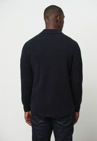 Suéter de punto azul marino con textura acanalada, cuello y mangas largas, usado sobre jeans de mezclilla oscuros, mostrado desde la parte de atrás.