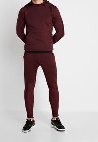 Bordeaux Nike hættetrøje med et sort logo og sorte detaljer, parret med matchende tætsiddende bukser med teksturerede paneler og en glat overflade.