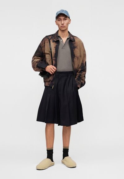 Άνδρας που φοράει μπλε καπέλο, καφέ και μαύρο μπουφάν, γκρι πόλο, μαύρη πλισέ φούστα, μαύρες κάλτσες, μπεζ παντόφλες και κρατάει μια μικρή μαύρη τσάντα.