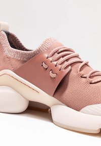 Różowe dziane sneakersy mają teksturowaną cholewkę, elastyczne sznurówki oraz brązowy skórzany akcent. Gruba, zaokrąglona biała podeszwa z subtelnymi detalami.