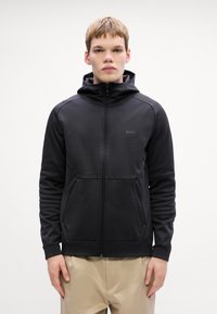 Črna jopica z zadrgo in kapuco, izdelana iz teksturiranega materiala, z dvema sprednjima žepoma na zadrgo in rebrastimi manšeti. Ima diskretno logotip znamke na prsih.