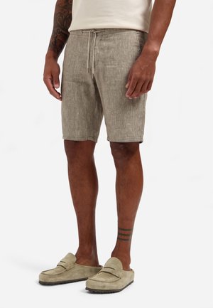 Mann trägt beige Leinen-Shorts mit Kordelzug, beige Loafers, tätowierte Arme und Unterschenkel, steht vor weißem Hintergrund.